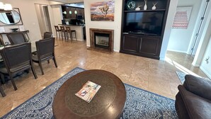 A living room with a fireplace, a TV, and a dining area with chairs and a kitchen in the background.