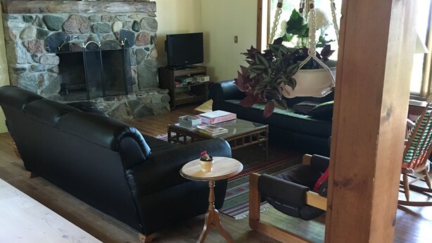 Ein gemütliches Wohnzimmer mit Kamin aus Stein, einer schwarzen Ledersofa, einem Holztisch und einer hängenden Makrameepflanze.