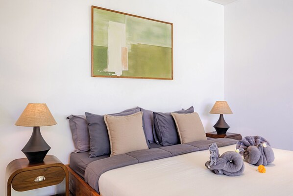 Un dormitorio ordenado con una cama, almohadas, dos mesitas de noche con lámparas, una mesita auxiliar de madera y un cuadro abstracto enmarcado en la pared.