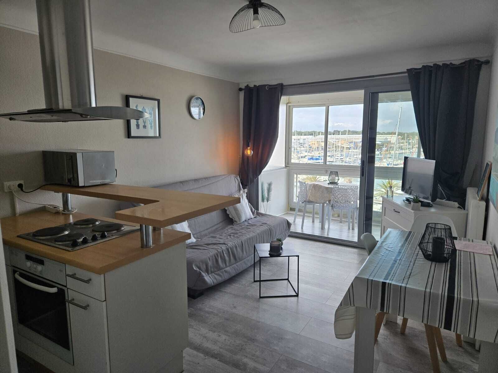 A modern apartment with a kitchenette, a sofa, a dining table, and a balcony with a view of the marina.