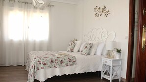 Een slaapkamer met een bed, witte gordijnen, een nachtkastje met een lamp en een decoratief wandobject.