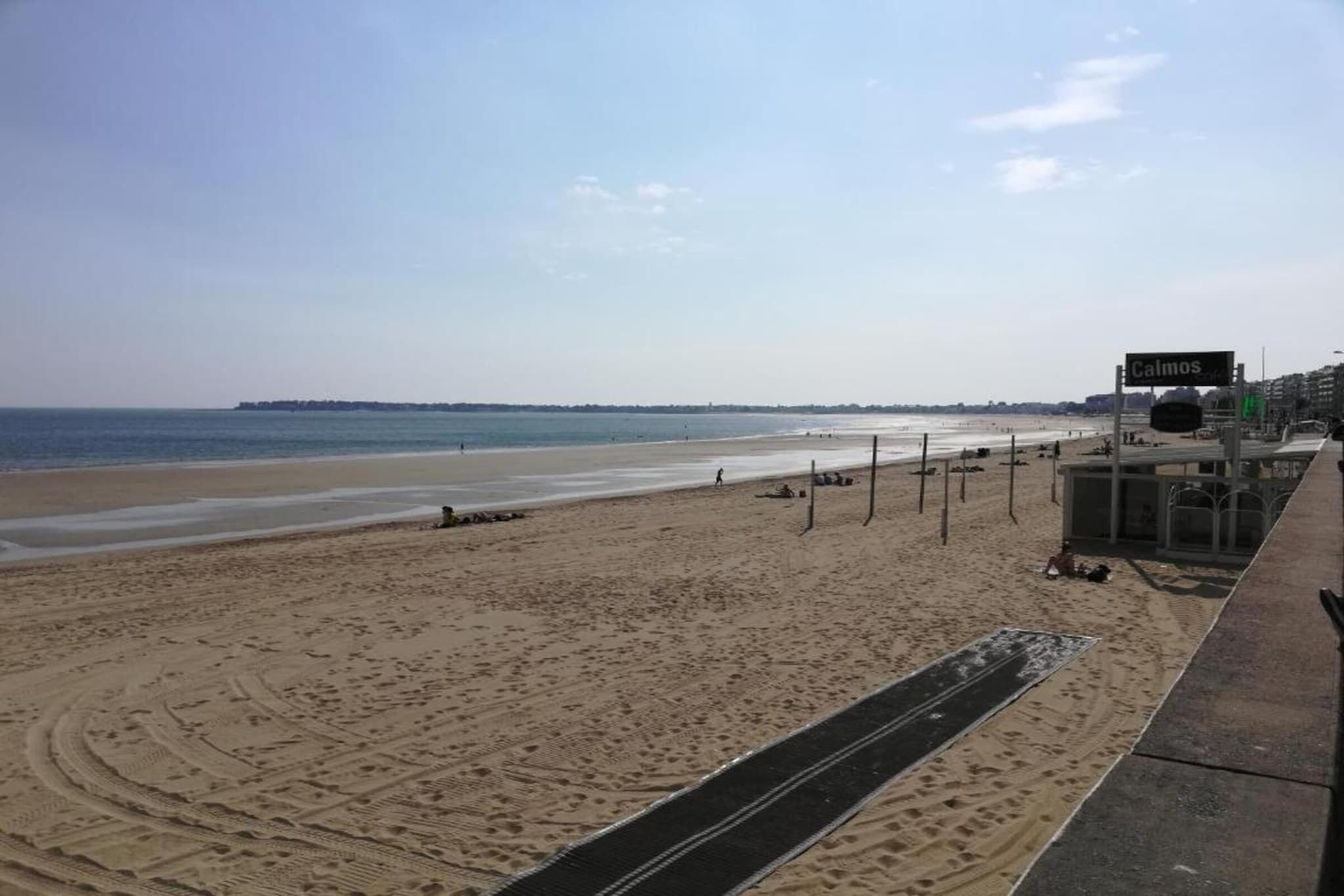 A beach with a clear sky, a sign reading 'Calmos', and a paved walkway.