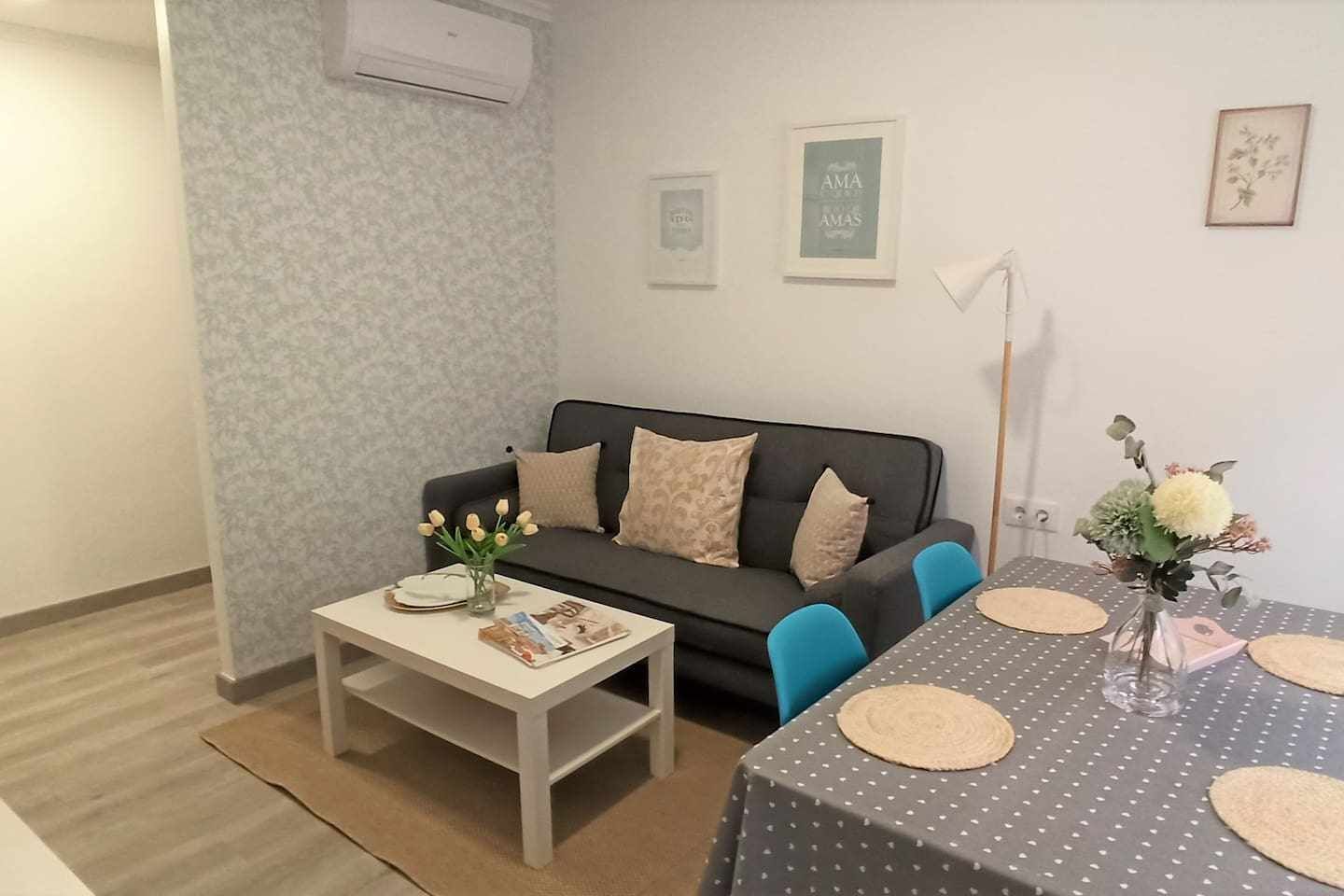 A living room with a sofa, a coffee table, a dining table with chairs, and a small vase with flowers.
