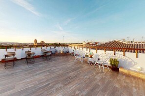 Une terrasse sur le toit, dotée d’un plancher en bois, de chaises et de tables en métal, et entourée de murs blancs et de plantes en pot.