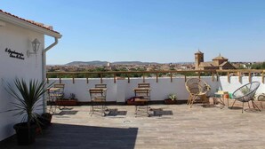 A rooftop terrace with wooden furniture, potted plants, and a view of a town with historical buildings. - Apartment amb Balcó (Consuegra)