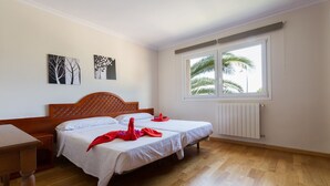 A bedroom with two beds, a wooden headboard, a window with a view of palm trees, and a radiator.