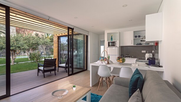 Ein modernes Wohnzimmer mit Sofa, Couchtisch und Essbereich mit Stühlen. Es gibt eine Küche mit weißen Schränken und einer Mikrowelle. Der Raum hat große Glasfenster, die auf eine Terrasse mit Blick auf Bäume und einen Rasen führen.
