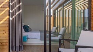 Ein modernes Zimmer mit einem Bett, einem kleinen Tisch und einem Stuhl. Eine Glastür öffnet sich zu einem Außenbereich mit Tisch und Stühlen.