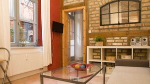 A living room with a glass coffee table, a sofa, a fireplace with a stone wall, a television mounted on the wall, and a window with a view of the outside. - Apartment lower deck with balcony - Gohr apartments (Stralsund)