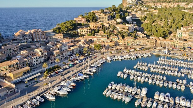 Uma cidade costeira com um porto de embarcações, rodeada por edifícios residenciais e uma colina com uma fortaleza.
