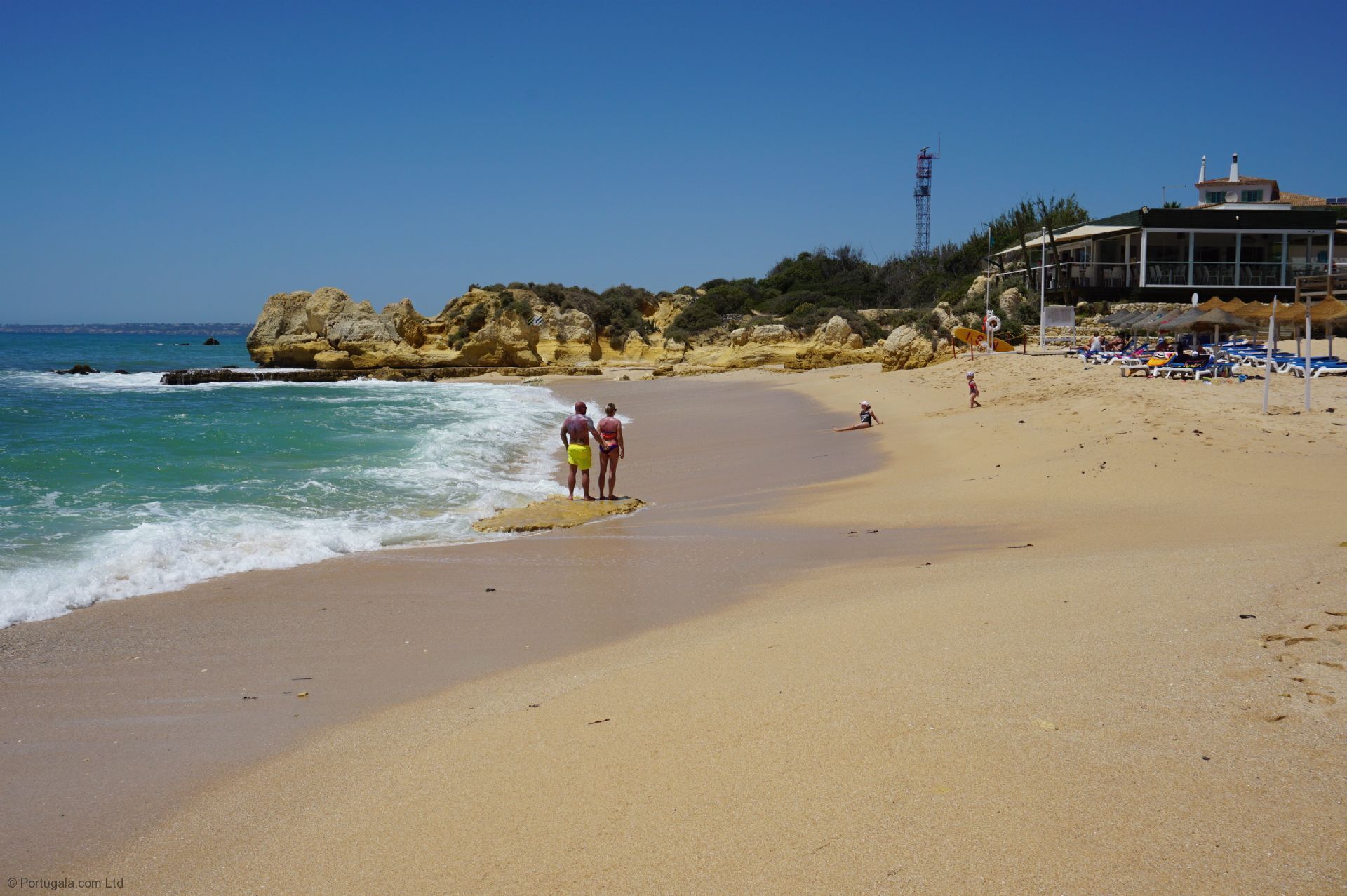 A beach with clear skies, rocky cliffs, and a building with a balcony overlooking the sea.