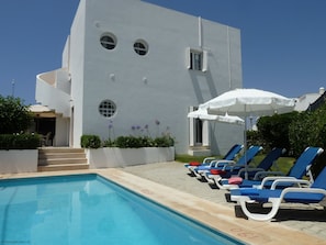 Un área de piscina con sillas de descanso y sombrillas, un edificio blanco con ventanas circulares y un cielo azul despejado.