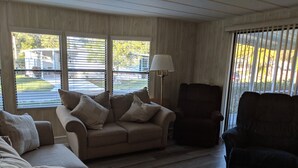 A living room with a beige sofa, a dark armchair, and a lamp. There are large windows with blinds, and a view of a parking lot and buildings outside. - A few short Miles from siesta key (Sarasota)