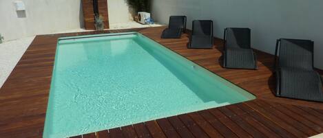 A rectangular swimming pool with a wooden deck, surrounded by lounge chairs and a white wall.