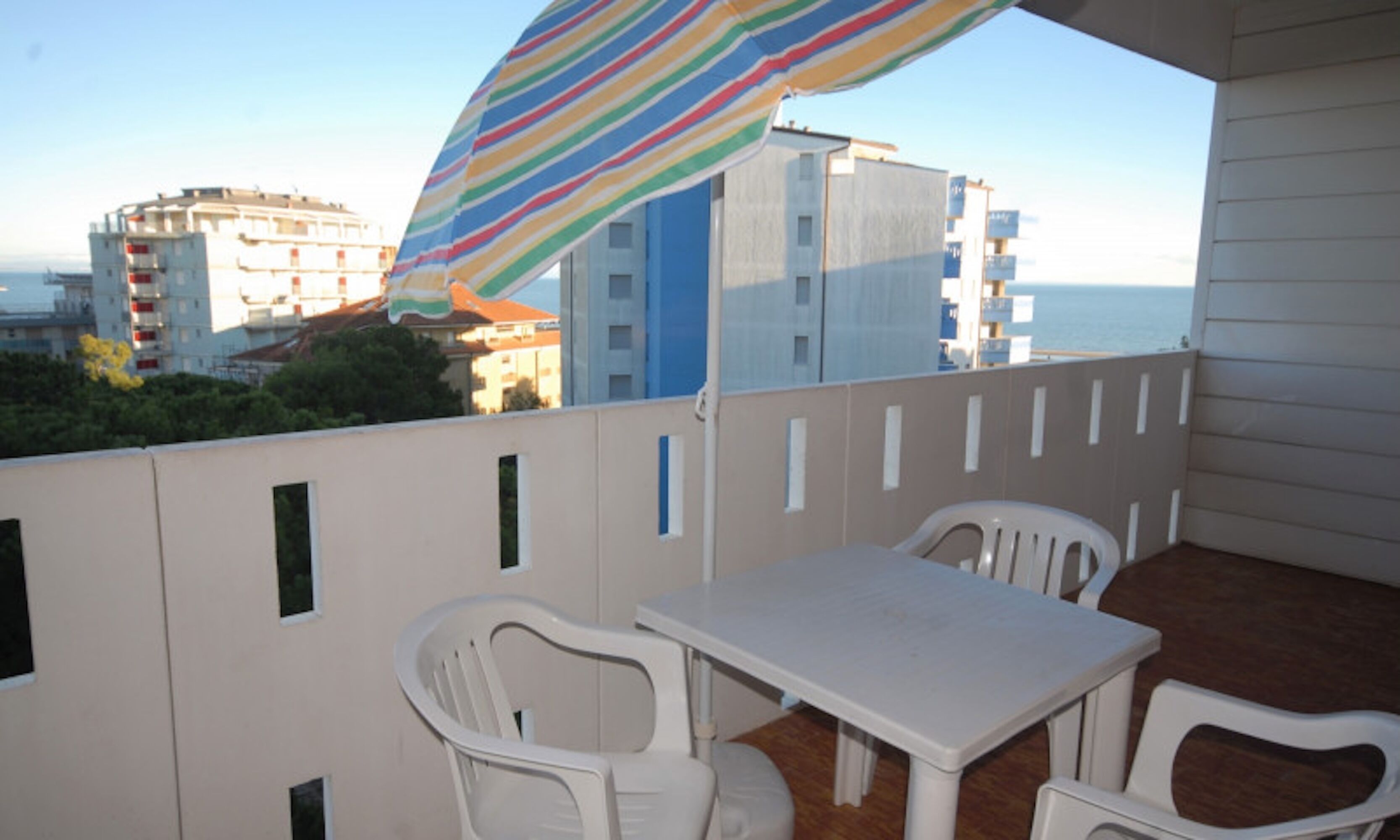 A balcony with a white table and chairs, a striped umbrella, and a view of buildings and the sea.