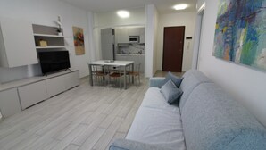 A modern living room with a grey sofa, a TV on a white cabinet, a dining table with chairs, and a kitchen in the background. - Bright flat with sea view (Lignano Sabbiadoro)