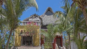A tropical resort with thatched-roof buildings, a white fountain, and palm trees.