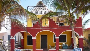 A beachside building with a red and yellow facade, white arches, and a thatched roof. There are white lounge chairs and a table with chairs in front.