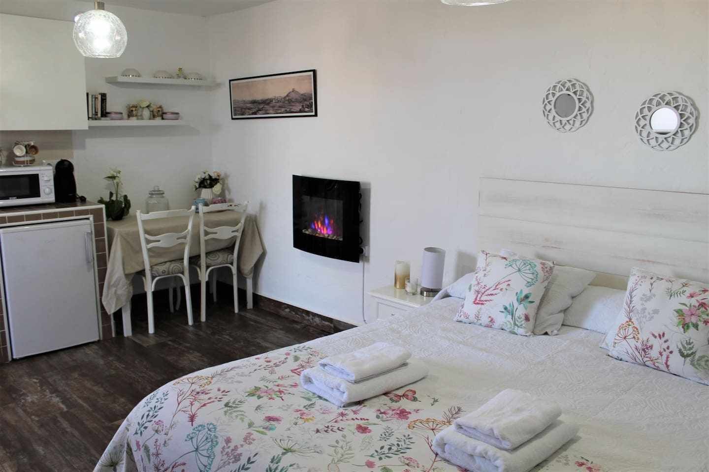 A compact hotel room with a bed, a small kitchenette, a dining area, and a fireplace.