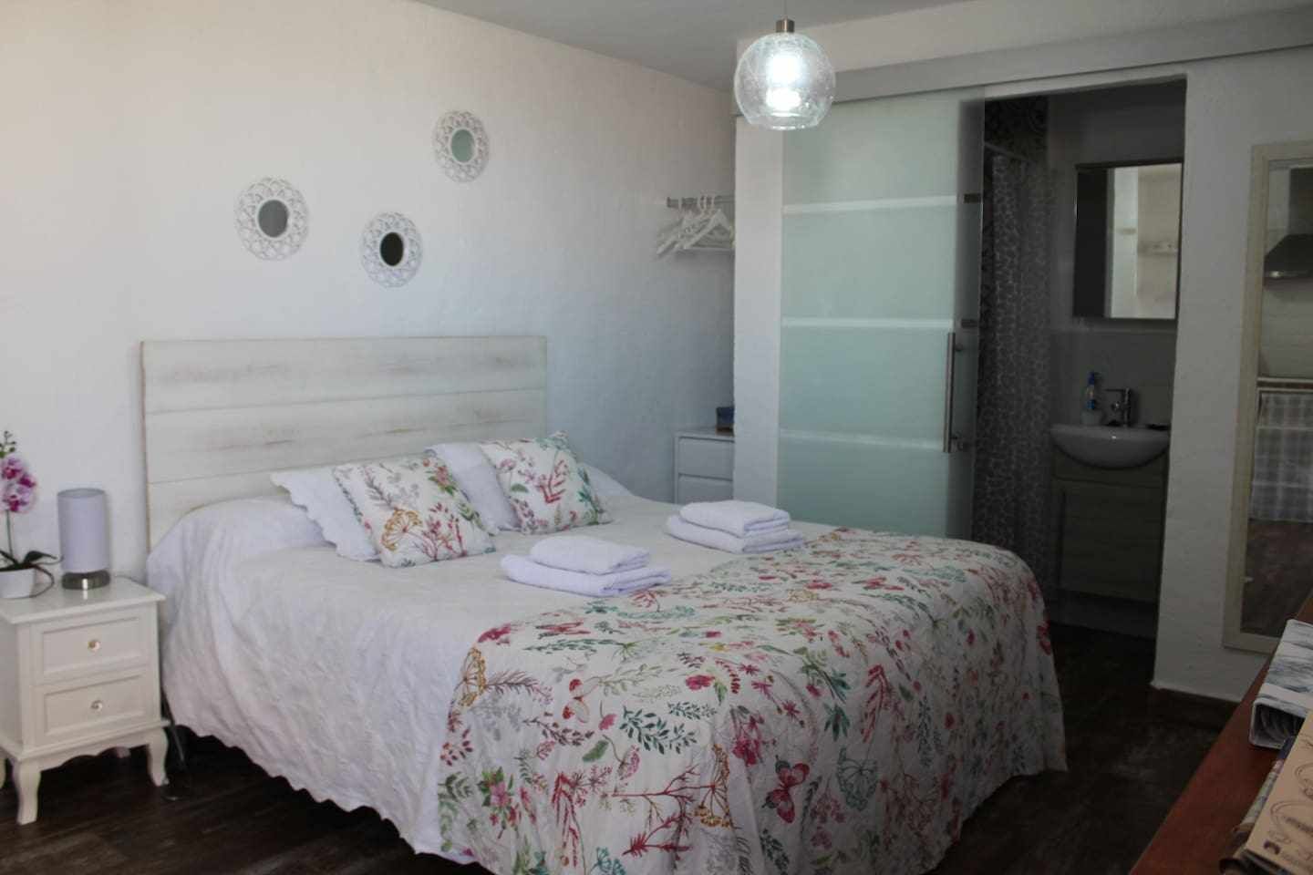 Un dormitorio con cama, mesita de noche, lámpara colgante y baño con lavabo y espejo.