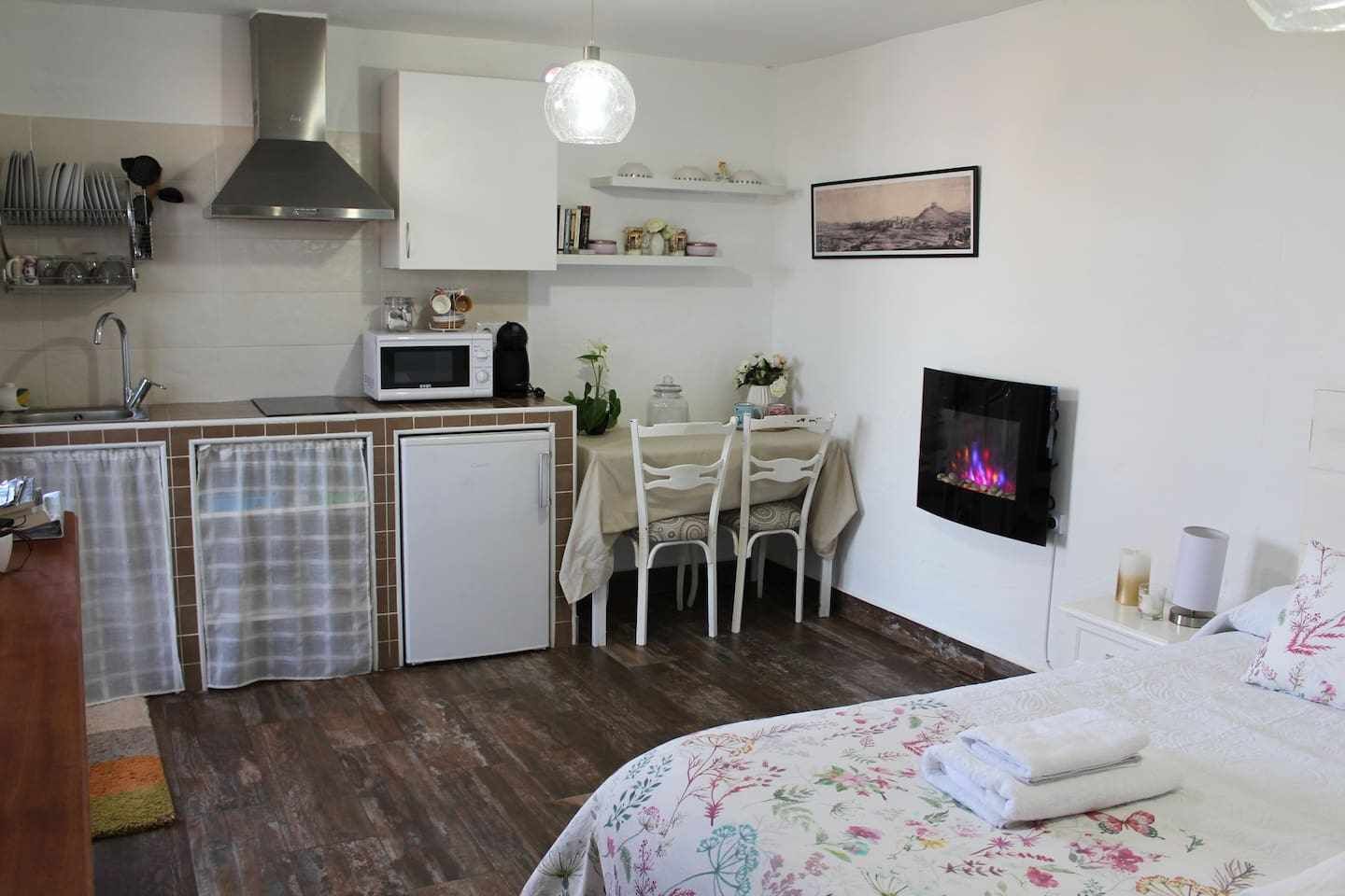 A compact kitchen with a microwave, sink, and a small refrigerator. There is a dining area with a table and chairs, and a fireplace on the wall.