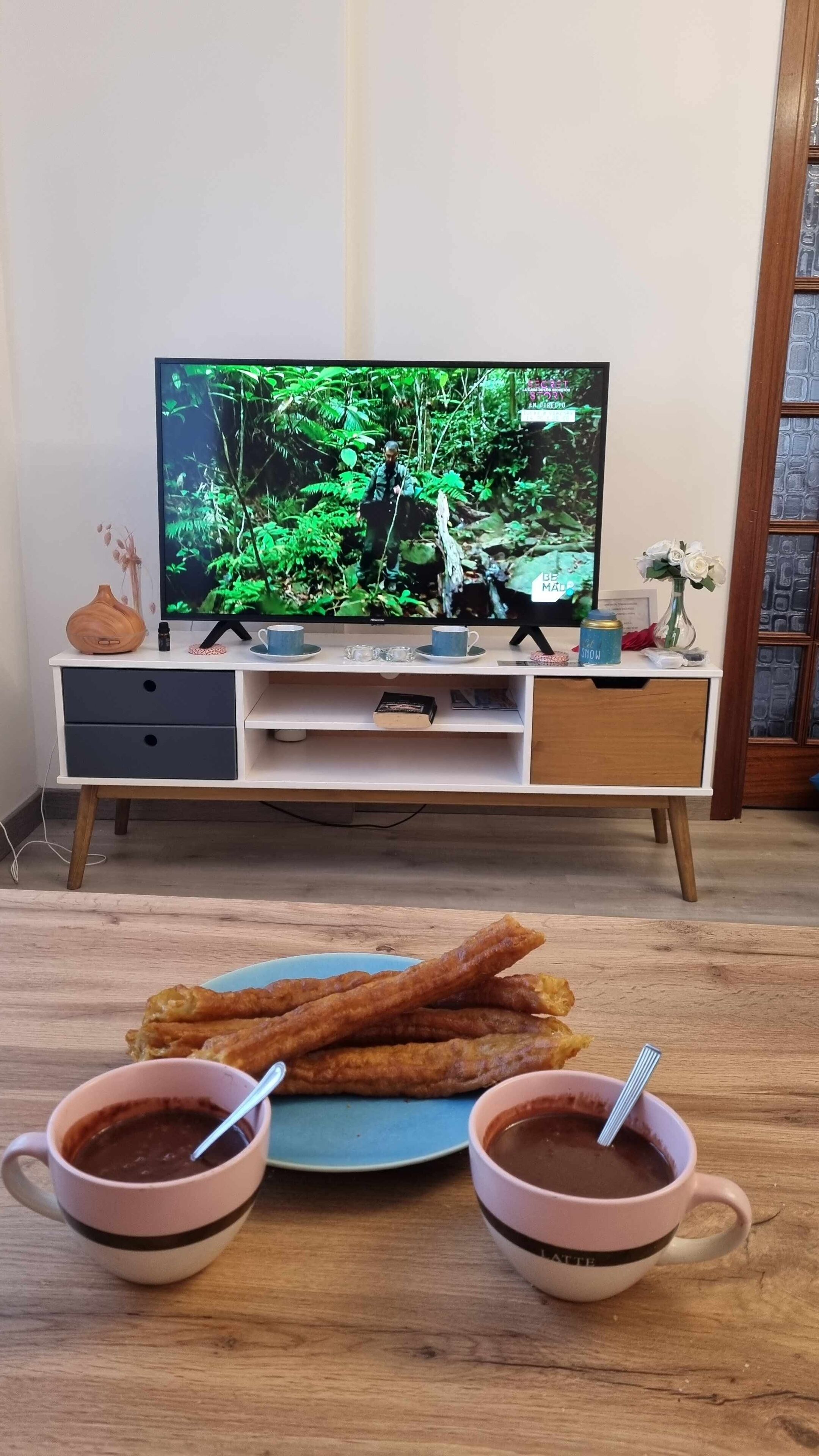 Una habitación con un televisor montado en la pared, una mesa de madera con un plato de churros y una taza de chocolate caliente, y un mueble pequeño con varios objetos.