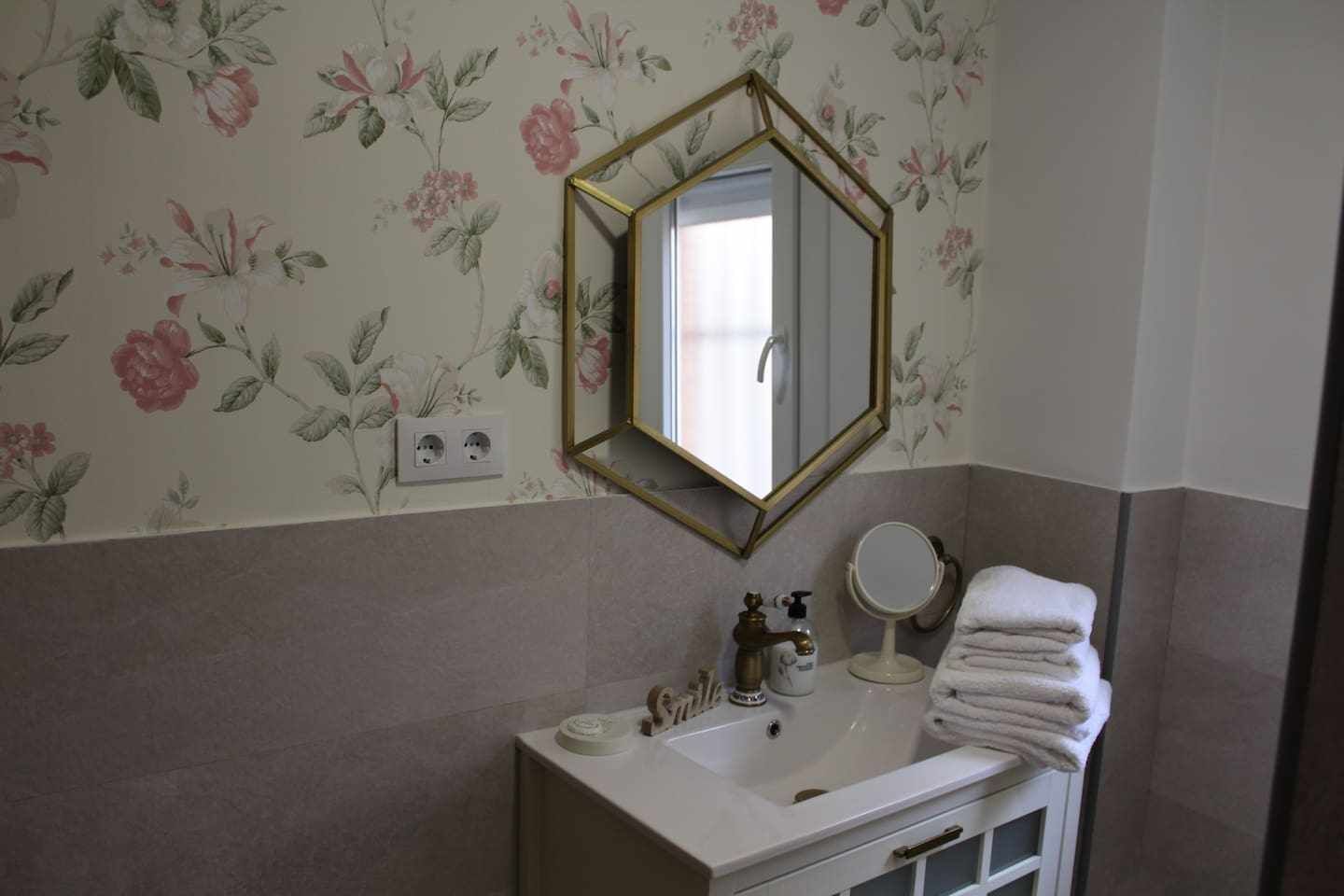 Un baño con papel tapiz floral, un espejo con marco dorado, un lavamanos con artículos de aseo y un perchero para toallas.