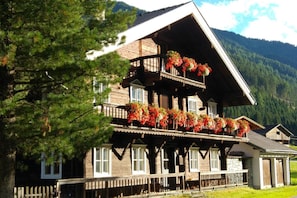 Ein traditionelles Holzchalet mit mehreren Balkonen, die mit Blumen geschmückt sind, umgeben von Wald und Bergen.