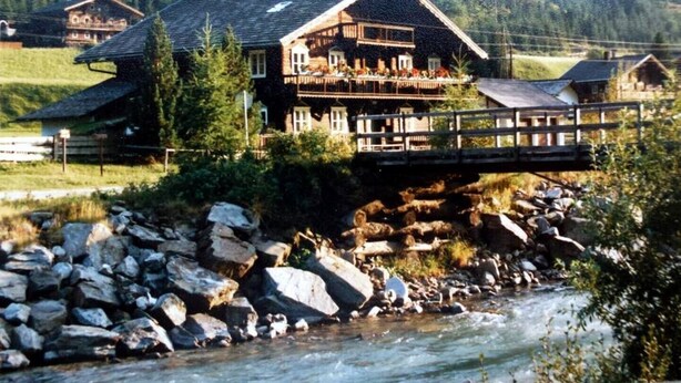 Ein Chalet mit Balkon, das auf einen Fluss, eine Holzbrücke und eine Berglandschaft blickt.