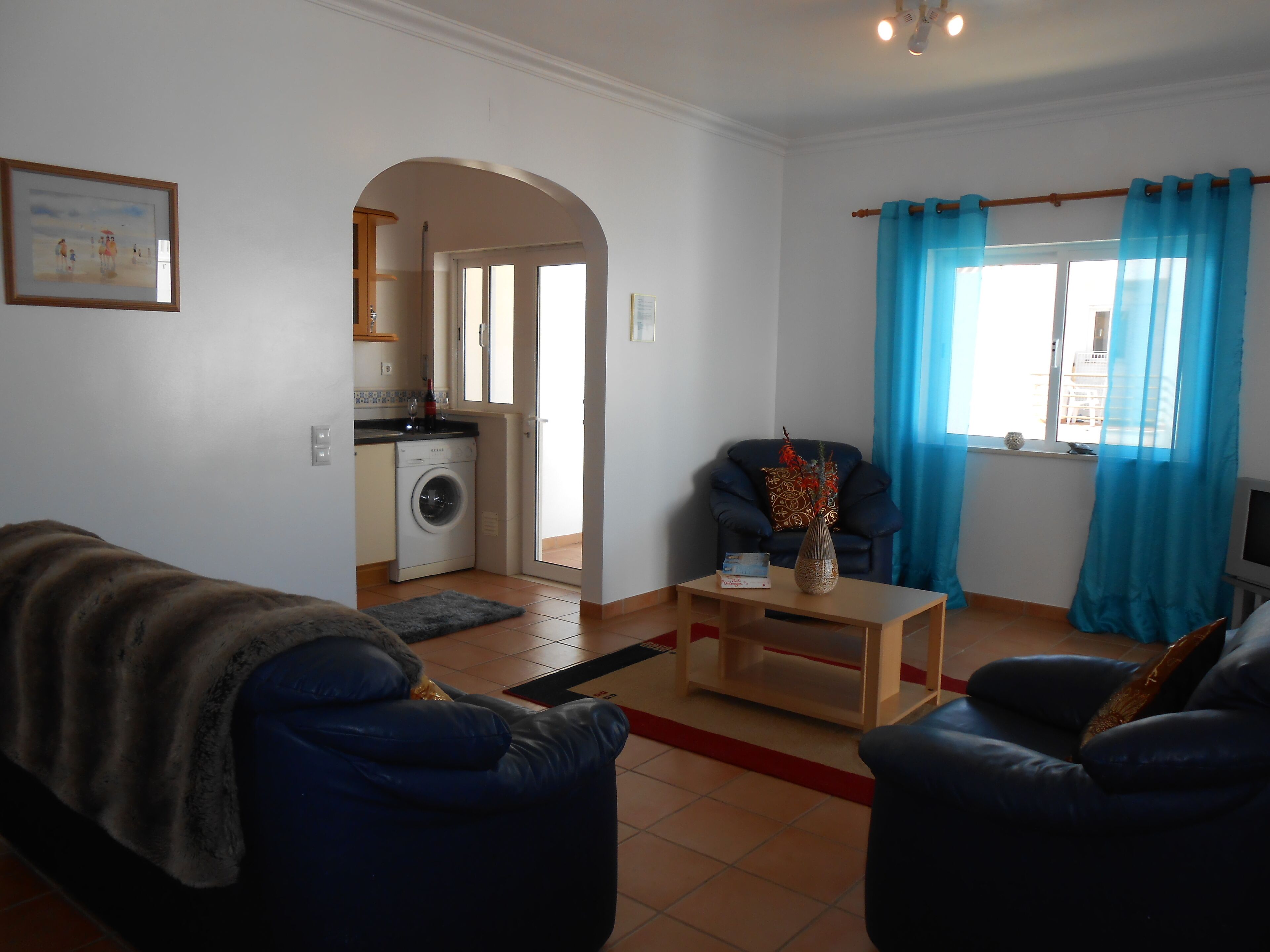A living room with a sofa, a coffee table, and a chair. There is a kitchen area visible through an archway.