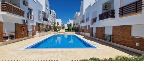 Una piscina rodeada de edificios modernos con balcones, un flotador salvavidas y un cielo azul despejado.