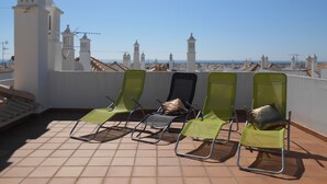 Une terrasse sur le toit, dotée de quatre fauteuils de jardin verts, d’un sol carrelé et offrant une vue sur des bâtiments blancs à multiples tours se découpant sur un ciel dégagé.