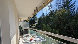 A balcony with a glass table set for dining, white chairs, and a view of trees and greenery. - Central Arcachon - Cozy T2 for 4 with Balcony, Parking, and Bike Storage (Arcachon)