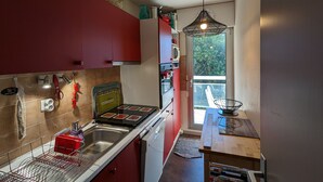 A compact kitchen with red cabinets, a stainless steel sink, and a small dining area with a wooden table. - Central Arcachon - Cozy T2 for 4 with Balcony, Parking, and Bike Storage (Arcachon)