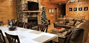Ein gemütliches Blockhaus-Interieur mit Kamin aus Stein, einem Weihnachtsbaum, einem Ledersofa und einem Esstisch für vier Personen.