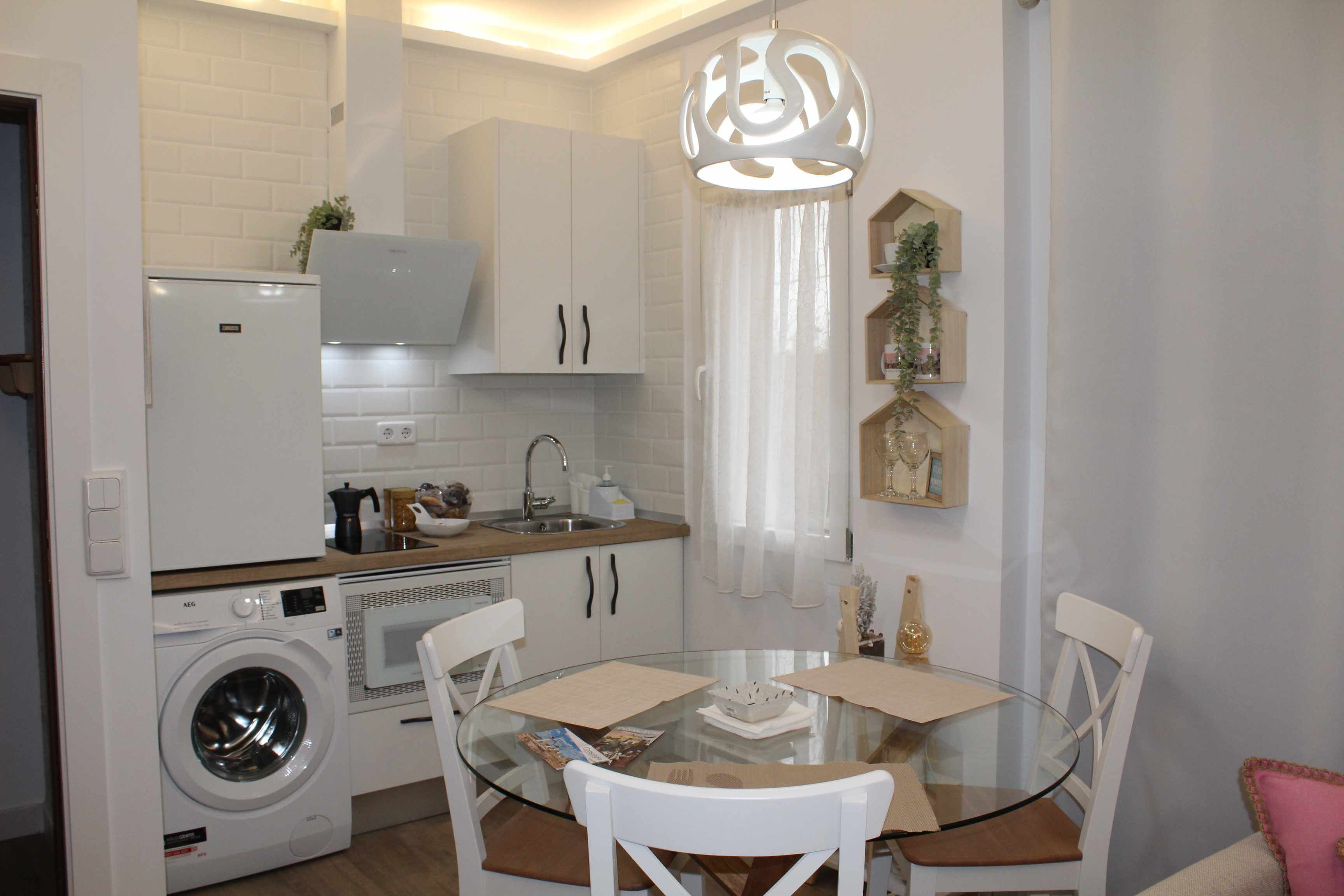 Cocina compacta con lavadora, microondas y comedor con mesa de vidrio y sillas blancas.