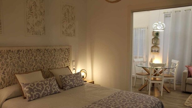 A bedroom with a large bed, patterned pillows, a bedside table with a lamp, and a view of a dining area with chairs and a table. - Apartment 2 Rooms (Consuegra)