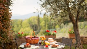 A round table with a basket, a cake, and plates of food, set outdoors with a view of trees and flowers. - Brunelleschi Apartment (Impruneta)
