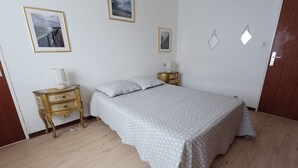 A bedroom with a bed, wooden floor, a nightstand with a lamp, and framed pictures on the wall. - 2-Bedroom Apartment with Balcony in Sables-d'Olonne (Les Sables-d'Olonne)