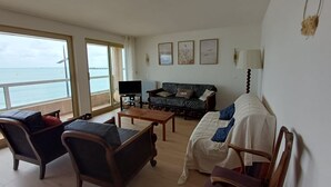 A living room with a sofa, chairs, a coffee table, and a TV. There are large windows with a view of the sea. - 2-Bedroom Apartment with Balcony in Sables-d'Olonne (Les Sables-d'Olonne)