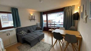 Una sala de estar moderna con un sofá gris, una mesa de comedor con sillas, una pequeña área de cocina y ventanas amplias con cortinas.
