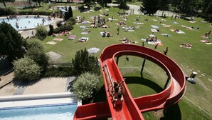 Un tobogán de agua en un parque con piscina y personas tomando el sol en el césped.