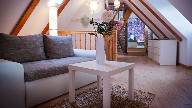 A cozy loft bedroom with a sloped ceiling, a wooden staircase, a white sofa, and a vase with flowers.