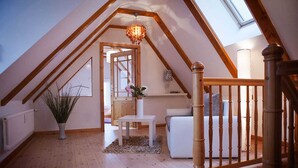 A cozy attic bedroom with a sloped ceiling, wooden beams, a skylight, a white sofa, a small table, and a potted plant.