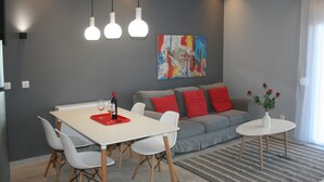 A modern living room with a dining table, white chairs, a grey sofa with red cushions, a colorful abstract painting on the wall, and a small white table with a vase of flowers. - Luxury Apartment, Tranquility, Sea Views, Breeze (Agios Gordios, Kerkira)