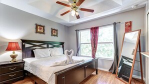 A bedroom with a wooden bed, a nightstand with a lamp, a ceiling fan, a window with curtains, and a mirror.