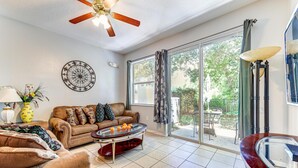 A spacious living room with a large window, a ceiling fan, a round wall clock, and a glass coffee table.