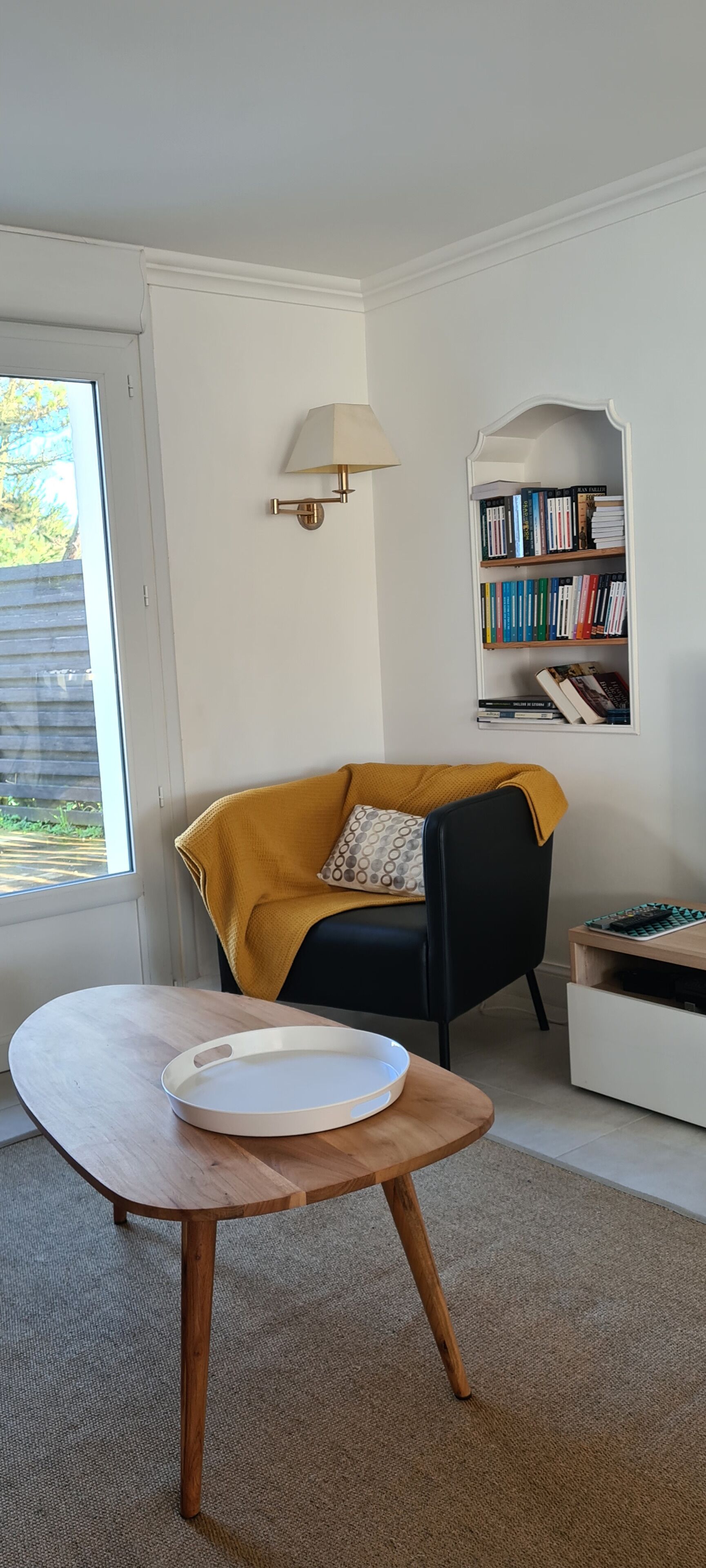 Une pièce avec une table en bois, une chaise noire agrémentée d’un coussin jaune moutarde, une bibliothèque et une lampe.