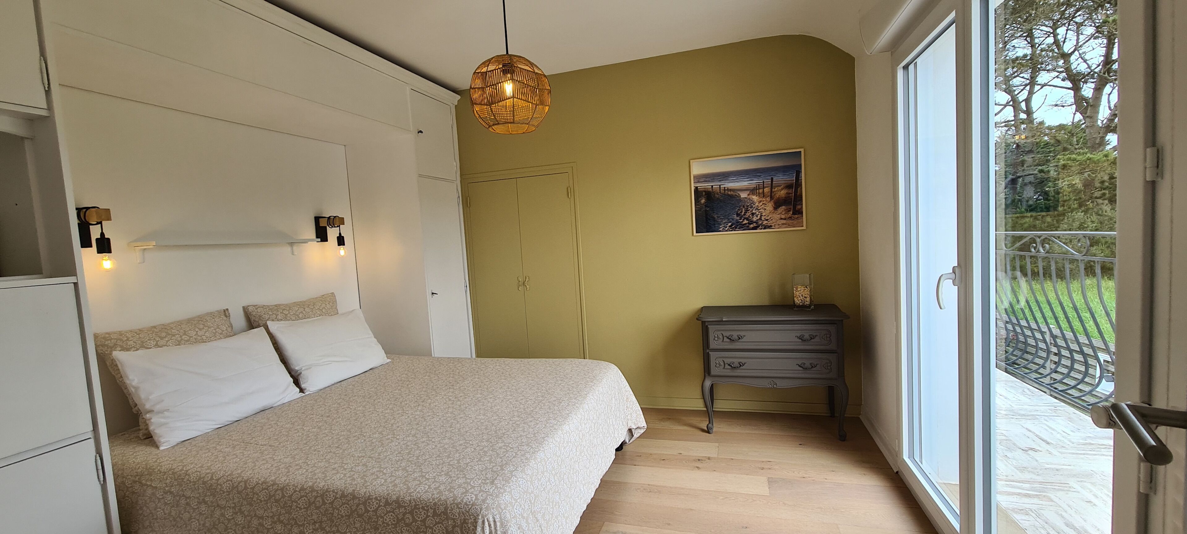 Ein Schlafzimmer mit Bett, Nachttischlampen, einem Schminktisch, einem Gemälde und Blick ins Freie.