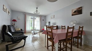 Ein Essbereich mit einem Tisch, gedeckt für vier Personen, umgeben von Holzsesseln. Ein schwarzer Sessel mit Kissen und eine rote Lampe sind links zu sehen. Über dem Tisch ist ein Deckenventilator angebracht.